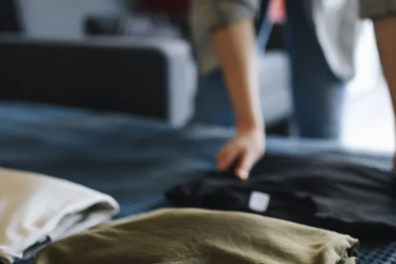 Plain T-shirts folded on a bed, in cream, khaki and black, as a hand tidies them away