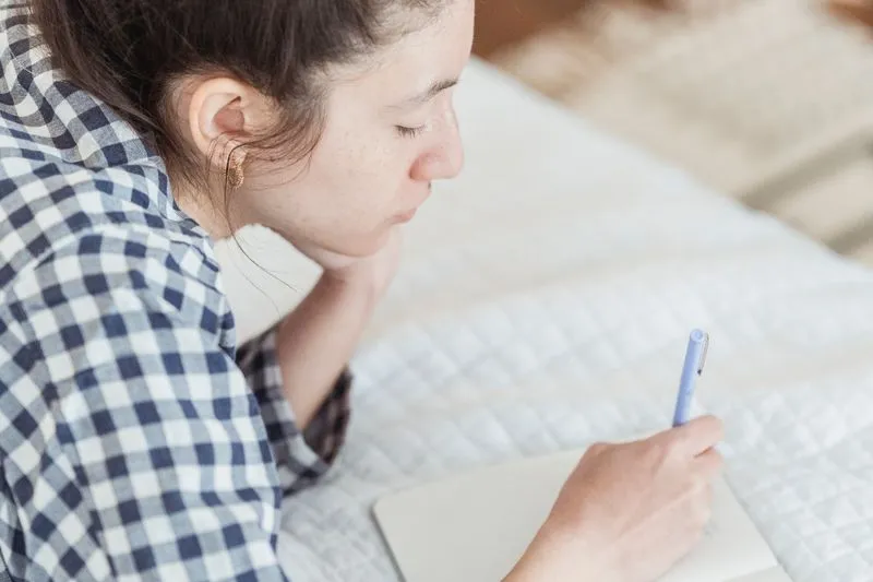 Woman in a checked shirt lying on her bed, writing in a notebook