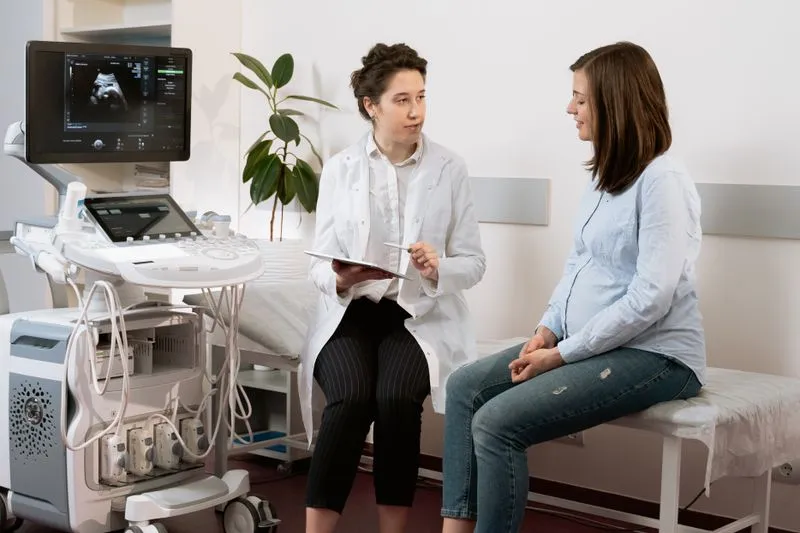 Pregnant woman sitting on the exam table, talking with a clinician taking notes in a bright consulting room with an ultrasound machine nearby