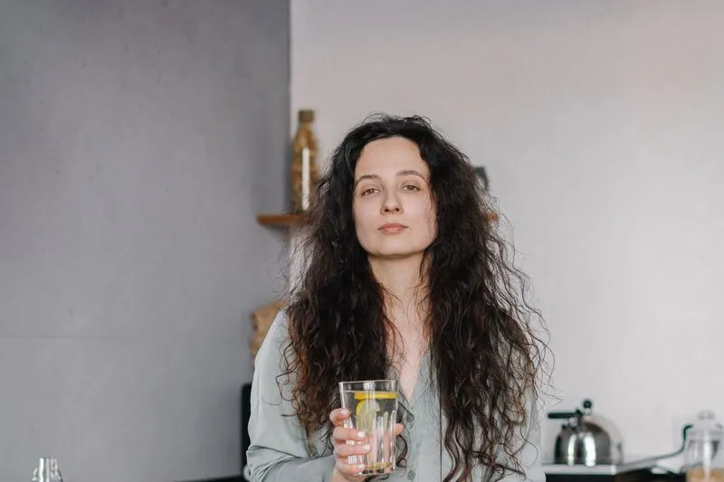Young woman with long curly hair, in pyjamas and looking tired, holding a glass of lemon water in her kitchen with a kettle behind her