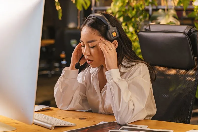 Woman taking a break at her desk, eyes closed and fingers pressed to her temples in front of her computer