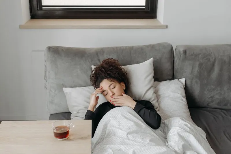 Young woman lying under a duvet on a grey sofa, eyes closed, one hand on her forehead and the other near her mouth, a cup of tea on the table beside her
