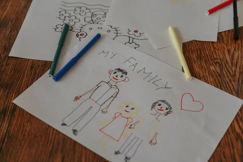 A child's drawing titled ‘My Family’, three felt-tip figures on a sheet of paper, surrounded by coloured pens on a wooden table