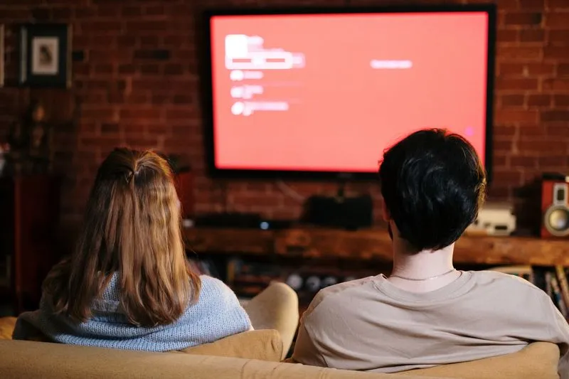 Two people seen from behind, sitting on a sofa in front of a large glowing television screen, in a brick-walled living room
