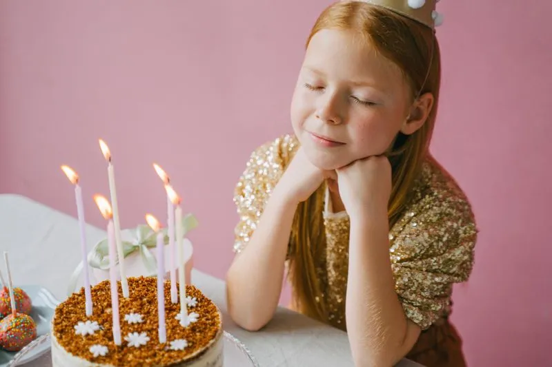 A girl wearing a gold crown sits with her eyes closed and her chin on her hands, in front of a birthday cake with lit candles, against a pink background