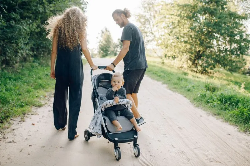 A couple walks along a tree-lined dirt path, pushing a toddler in a pushchair