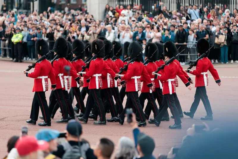 Guards in red tunics and bearskin hats marching past a crowd, near the gates of Buckingham Palace