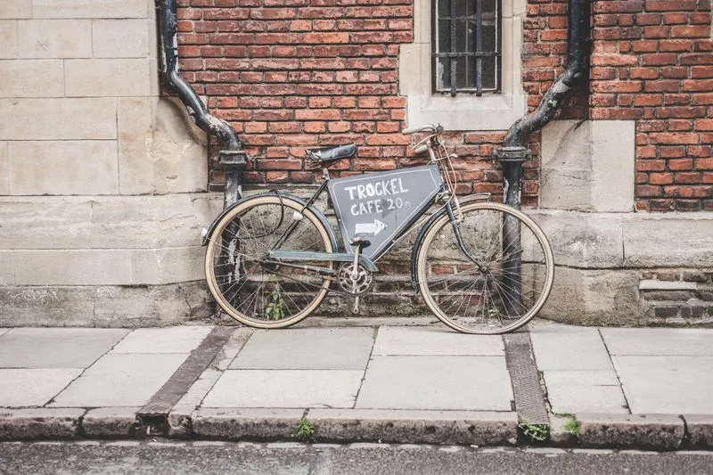 Old bicycle with a 'Trockel Café 20 m' sign on its frame, leaning against a brick wall