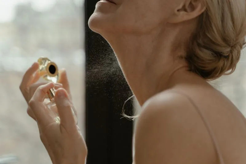 Woman spraying perfume near her neck by a bright window, the mist visible in the light