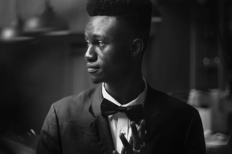 Black-and-white portrait of a young Black man in a dark suit jacket, white shirt and bow tie, looking to the side