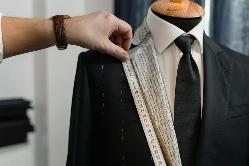 Tailor's hand taking measurements on a black suit jacket over a dressmaker's dummy already dressed in shirt and tie