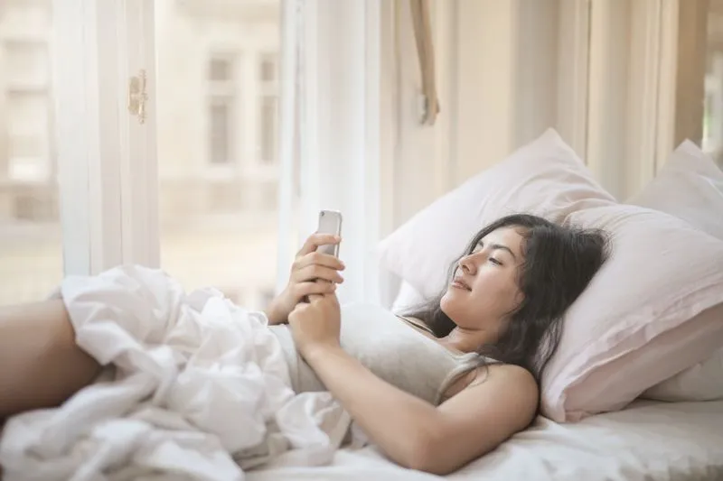 Young woman lying in bed early in the morning, looking at her phone screen