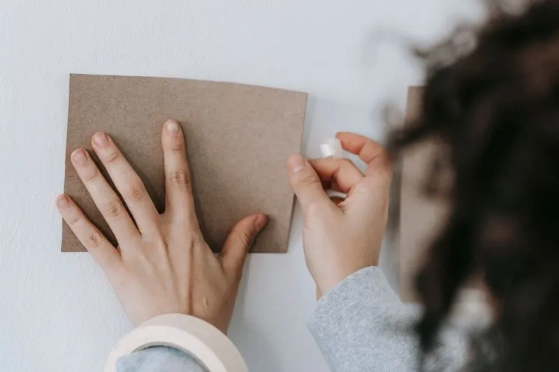 Hands fixing a square of kraft card to a white painted wall with adhesive tape, a roll of masking tape in the foreground