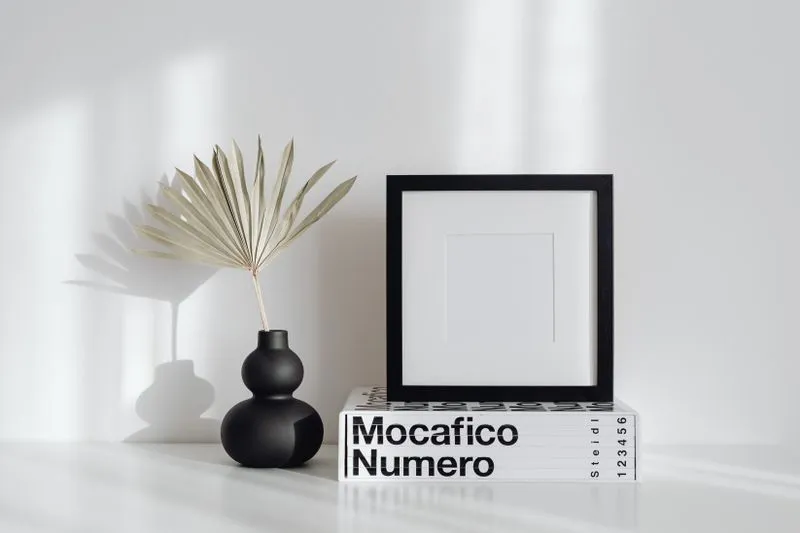 Black frame with a white mount resting on a large book against a white wall, next to a black vase holding a dried palm leaf