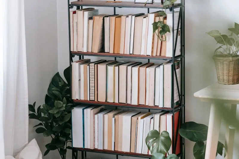 Black freestanding shelving unit filled with books, surrounded by potted green plants against a white wall