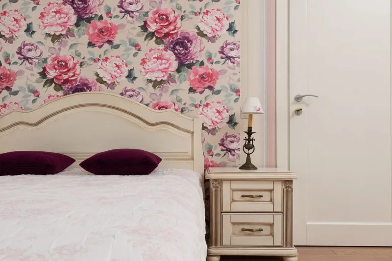 Large pink and purple floral wallpaper on the wall behind a cream headboard, with the neighbouring wall left white around the door