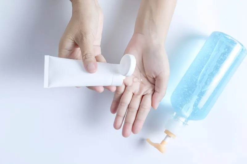 Hands squeezing a few drops of clear gel from a plain white tube, next to a pump bottle of pale blue gel