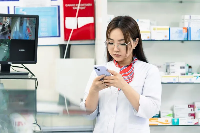 Young pharmacist in a white coat holding her phone in front of shelves of medicine boxes