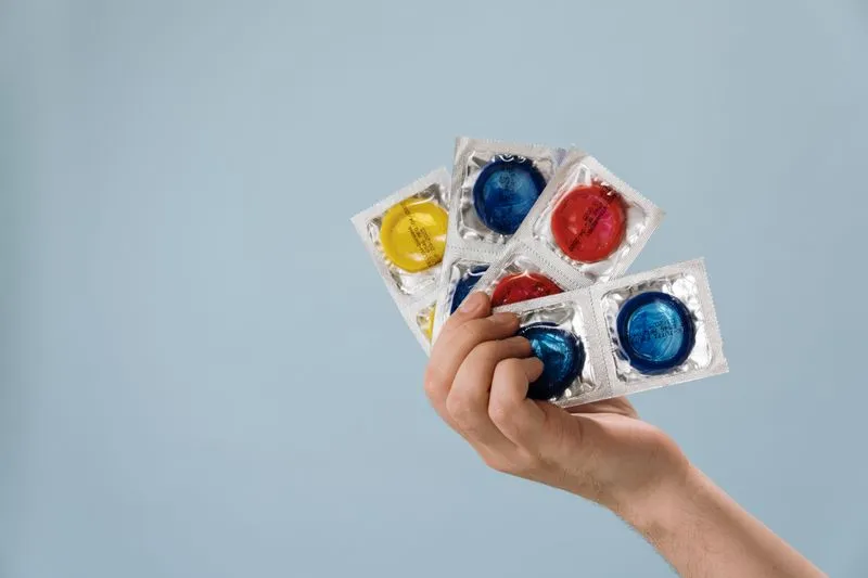 Hand holding a fan of wrapped condoms in yellow, blue and red against a pale blue background