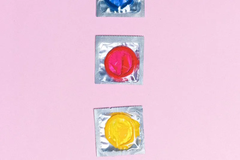 Three condom wrappers, blue, pink and yellow, lined up on a pink background
