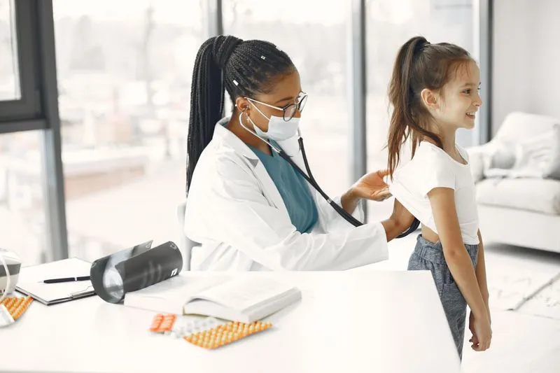 A paediatrician listens to a smiling young girl's back with a stethoscope in a clinic room