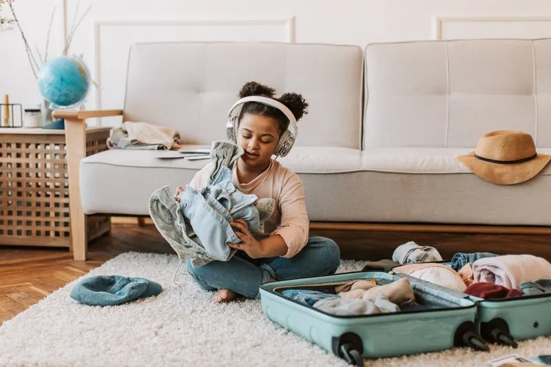 A girl sitting cross-legged with headphones on, folding clothes beside a half-packed open suitcase