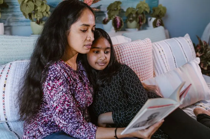 A mother and her daughter of about ten, snuggled up on a sofa, reading a book together