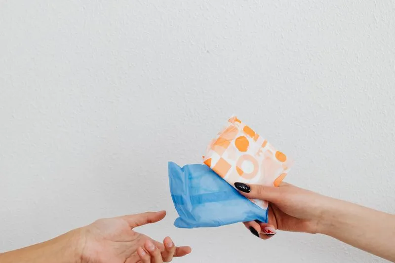 Close-up of two hands: one passing two individually wrapped sanitary pads to the other