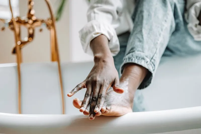 A hand smoothing cream onto the heel of a foot resting on the edge of a bath