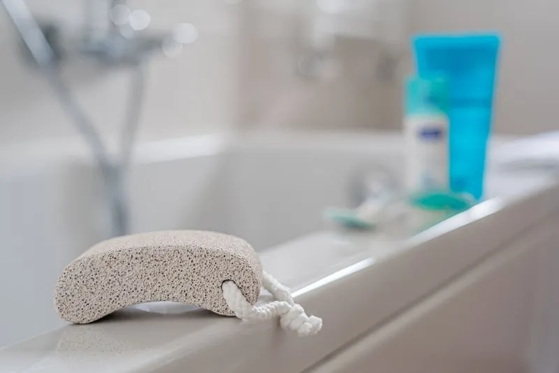 A grey pumice stone on the edge of a bath next to skincare products