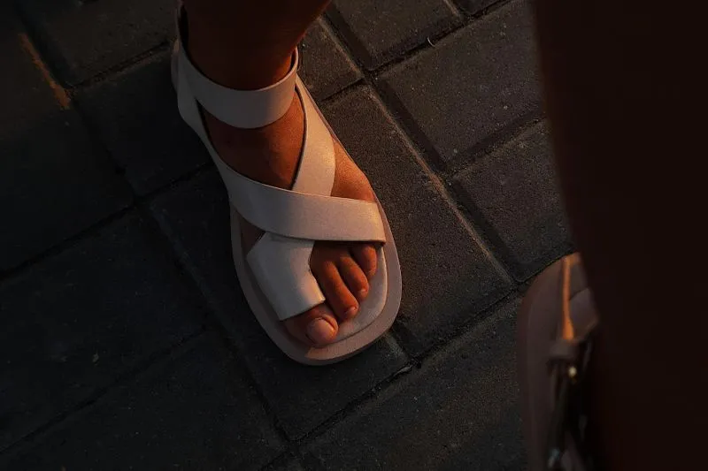 A woman's foot in a pale crossover-strap sandal, resting on paving stones lit by golden light