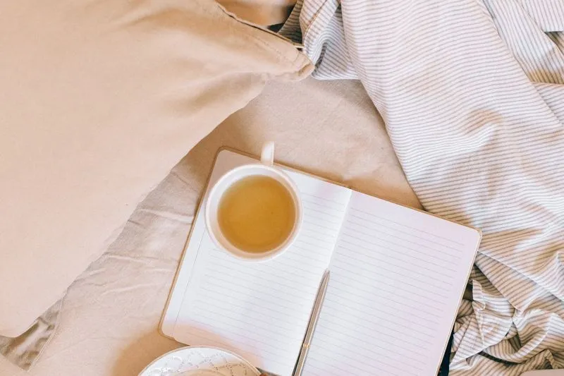 An open notebook with a pen, resting on a bed beside a mug of herbal tea