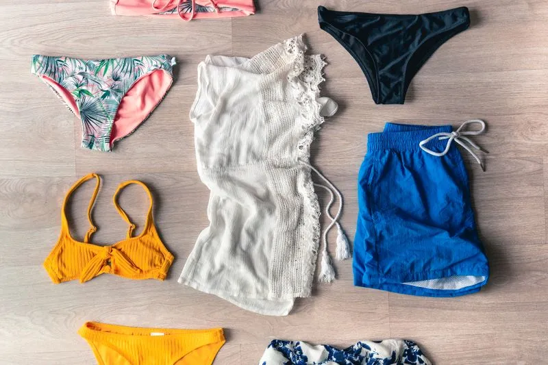 Bikini tops and bottoms in different colours and shapes, a lace cover-up and blue swim shorts, laid flat on a wooden floor