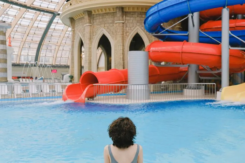 Woman seen from behind, grey swimsuit straps, facing the slides of an indoor pool