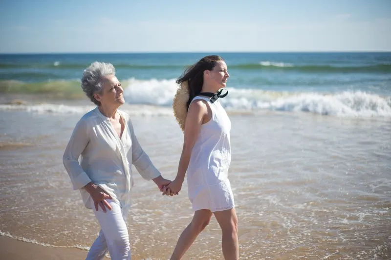 Dark-haired woman and a white-haired older woman walking hand in hand along the water's edge on a sunny beach