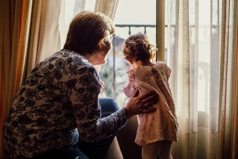 Older woman seen from behind, steadying a little girl standing at a window with pale net curtains