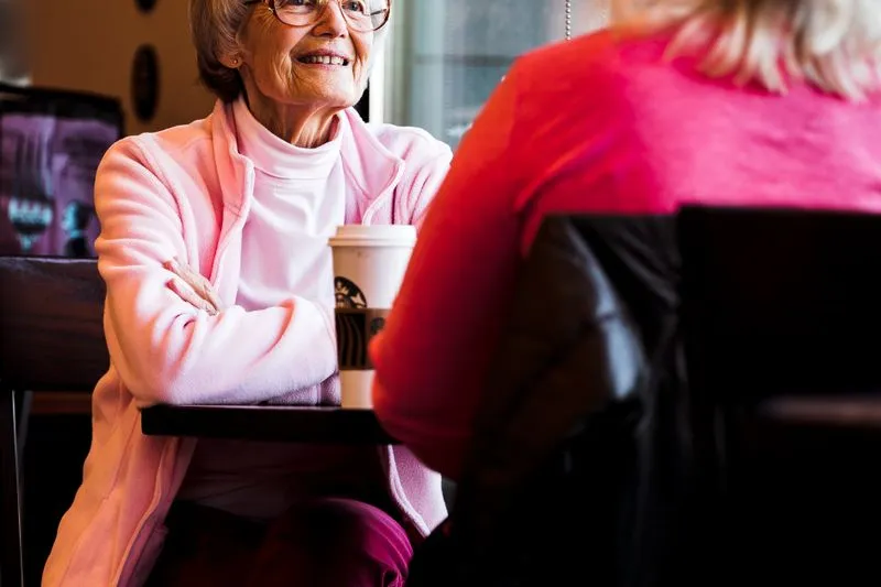 Smiling older woman at a café table, facing a woman seen from behind, a takeaway cup on the table