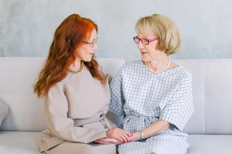 Mother with an ash-blonde bob and her red-haired adult daughter sitting side by side on a sofa, talking and holding hands