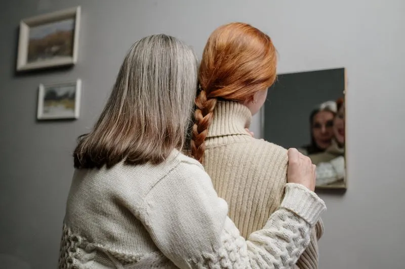 Grey-haired woman with her hand on the shoulder of her adult daughter, who wears a red plait, both seen from behind facing a mirror