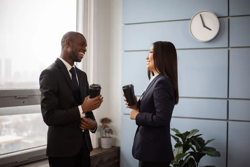 Two smiling colleagues chatting in an office, coffee cups in hand