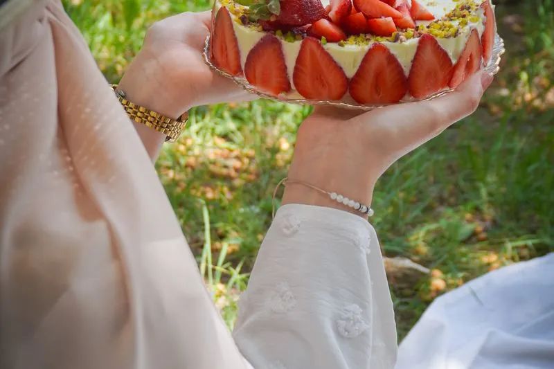 Whole fraisier held in both hands in a garden, halved strawberries pressed all round the side, topped with strawberries and chopped pistachios