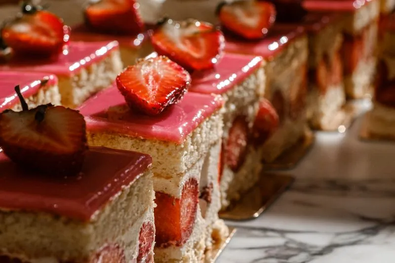 Square slices of fraisier lined up on a marble counter: génoise, cream and cut strawberries visible on the side, pink glaze and half a strawberry on top