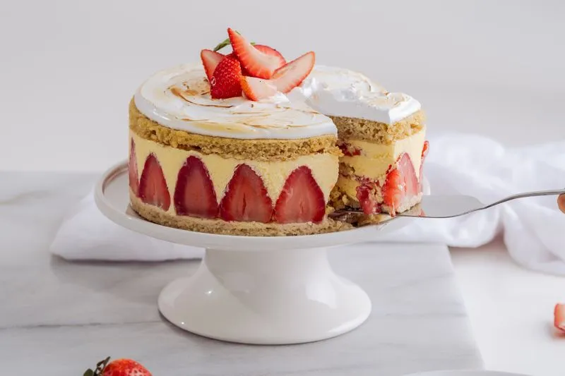 Fraisier on a white cake stand: halved strawberries lined up round the side, a torched meringue top with fresh strawberries, one slice being lifted out with a cake server