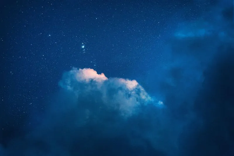 Star-filled deep blue night sky with a small glowing nebula above a dusk-lit cloud
