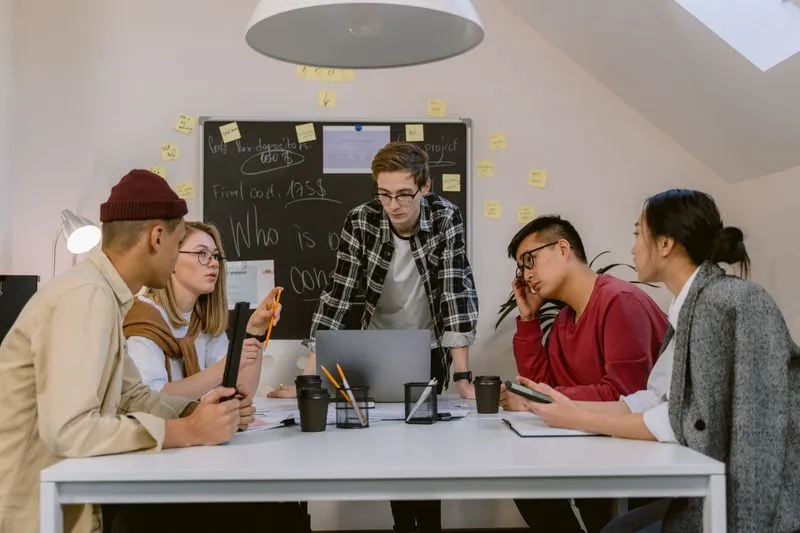 Young man presenting a project in front of a board covered in sticky notes, surrounded by colleagues seated around the table