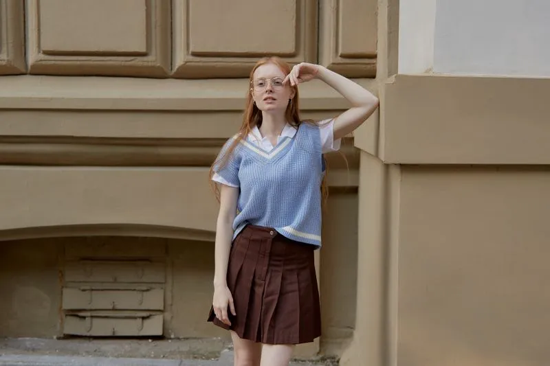 Young woman in a brown pleated skirt and a pale blue sleeveless V-neck jumper worn over a white shirt