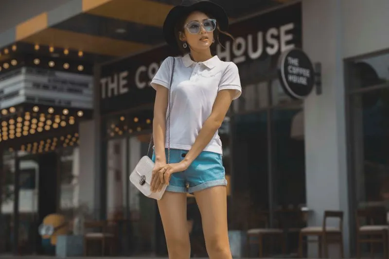 Young woman in a white polo shirt and denim shorts, with a black hat and sunglasses, in front of a café