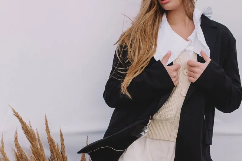Dark blazer open over a white shirt with a ruffled collar, a buttoned ecru knitted waistcoat and cream trousers
