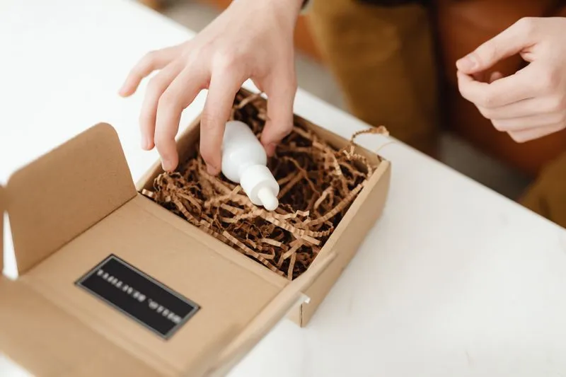 Hand lifting a small plain white dropper bottle, with no label, out of a cardboard parcel