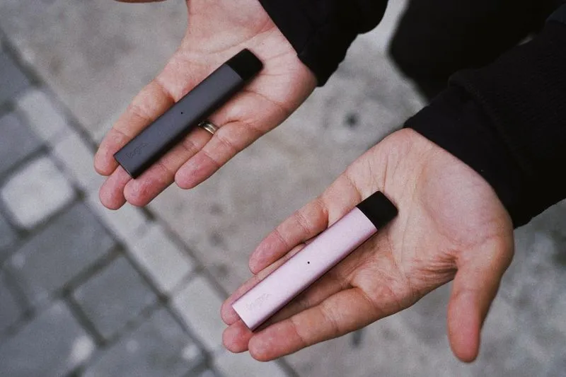 Two vape pens, one grey and one pink, resting in the palms of two hands